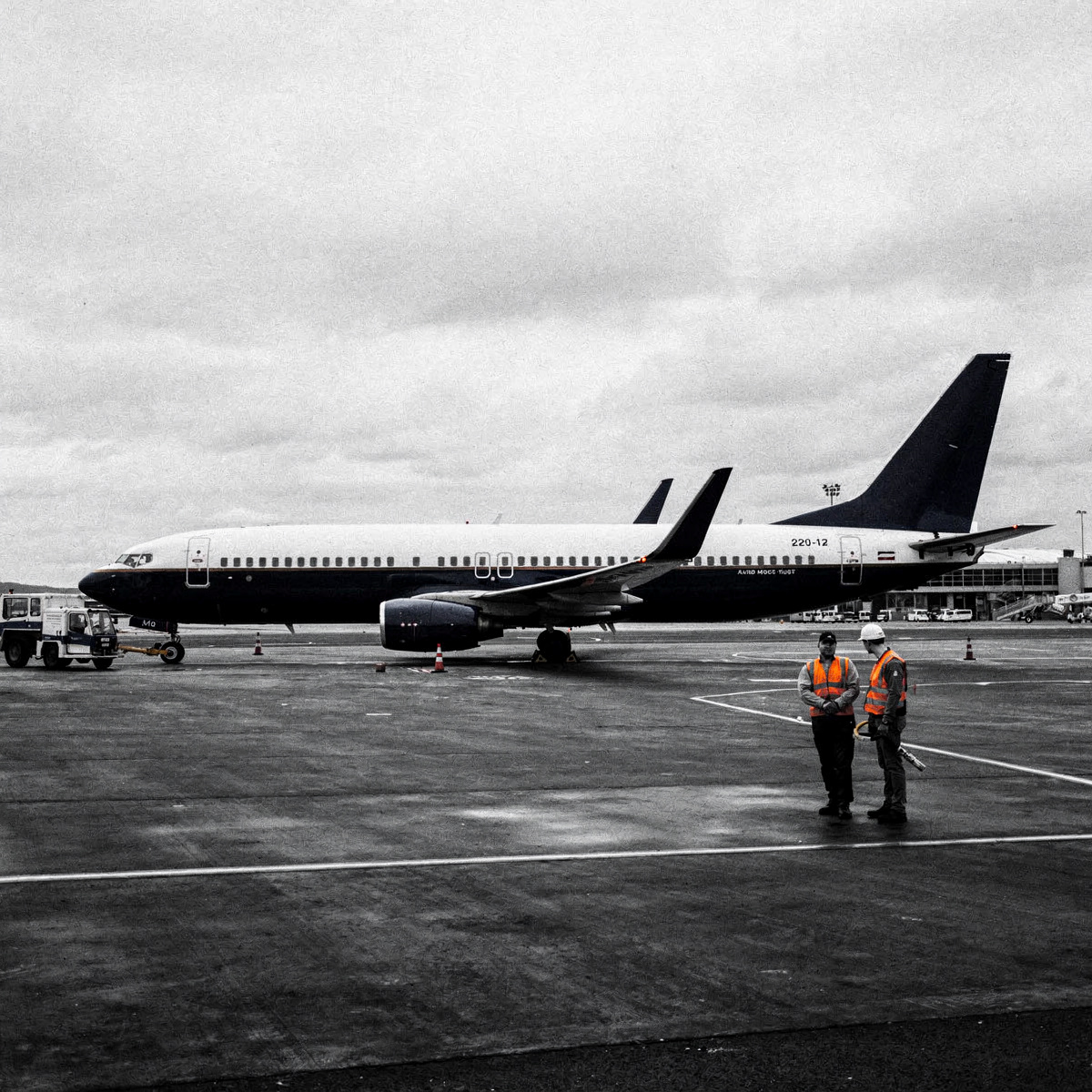 A Boeing 737-800 aircraft is parked at a remote stand on an airfield apron under an overcast sky. Ground equipment is working around it.