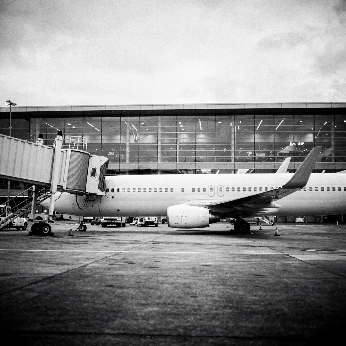 An unbranded Boeing 737-800 aircraft is parked at a gate under an overcast sky, with a jetbridge connected to its forward door.