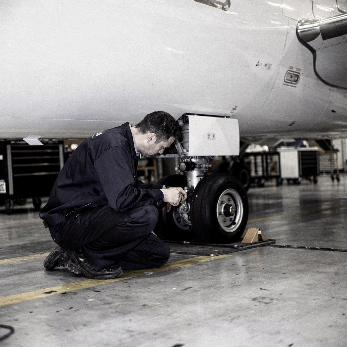 A maintenance worker in a dark uniform is crouched beside the landing gear of a white A320neo family aircraft on a hangar floor, inspecting a component.