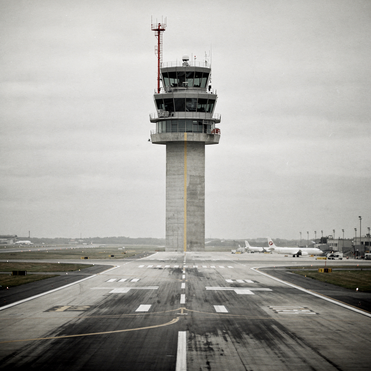 An airport control tower stands tall against an overcast sky, its structure dominating the frame, with a single runway stretching into the distance.