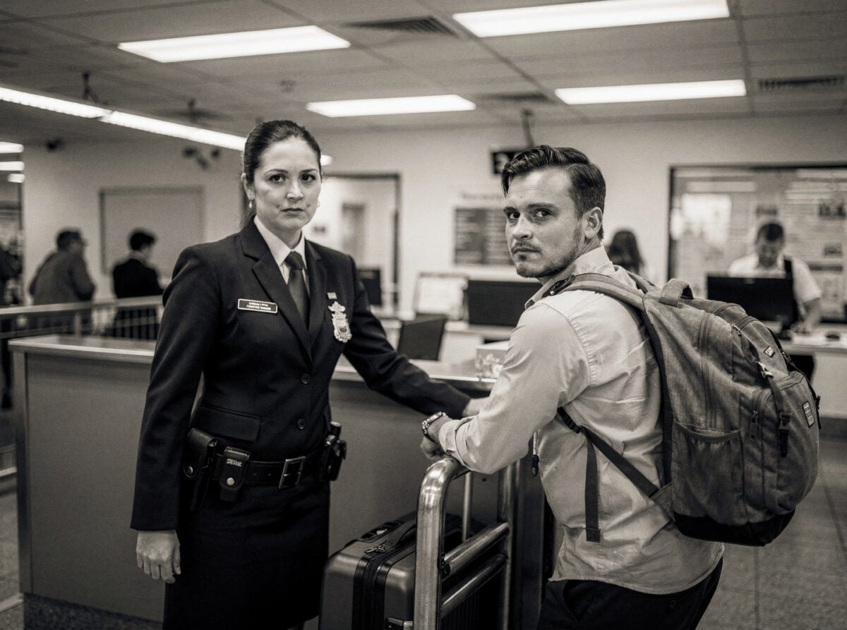 An Australian border control officer stands at a customs checkpoint, examining a passenger's luggage. It is daytime.