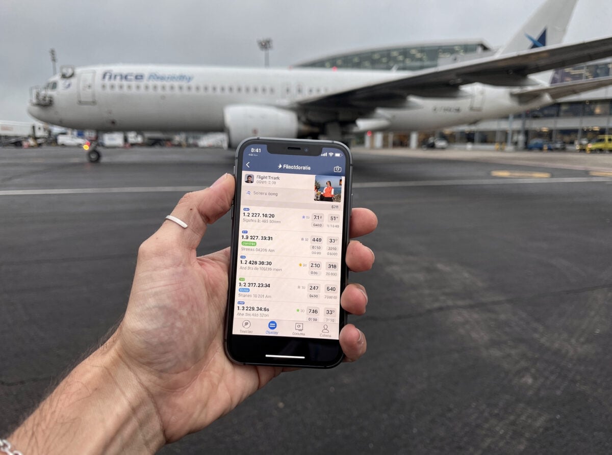 A single hand holding a smartphone, displaying a flight tracking application, with an aircraft partially visible in the background on an overcast day.