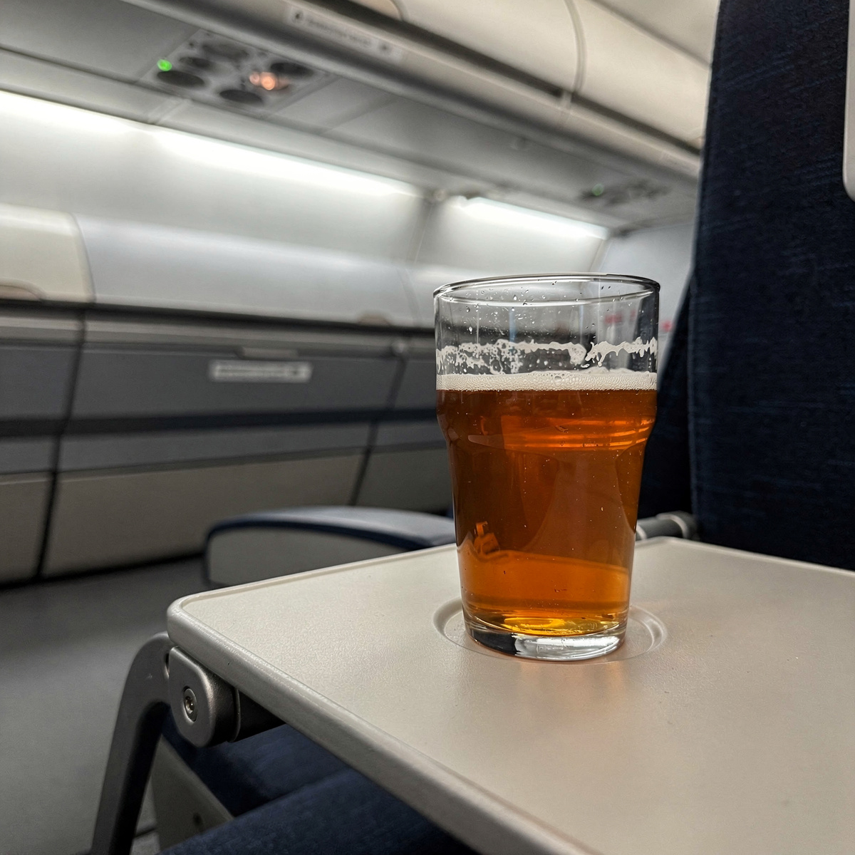 A single glass of amber-colored beer sits on a neutral-toned tray table in a commercial aircraft cabin, illuminated by cool white overhead lighting.