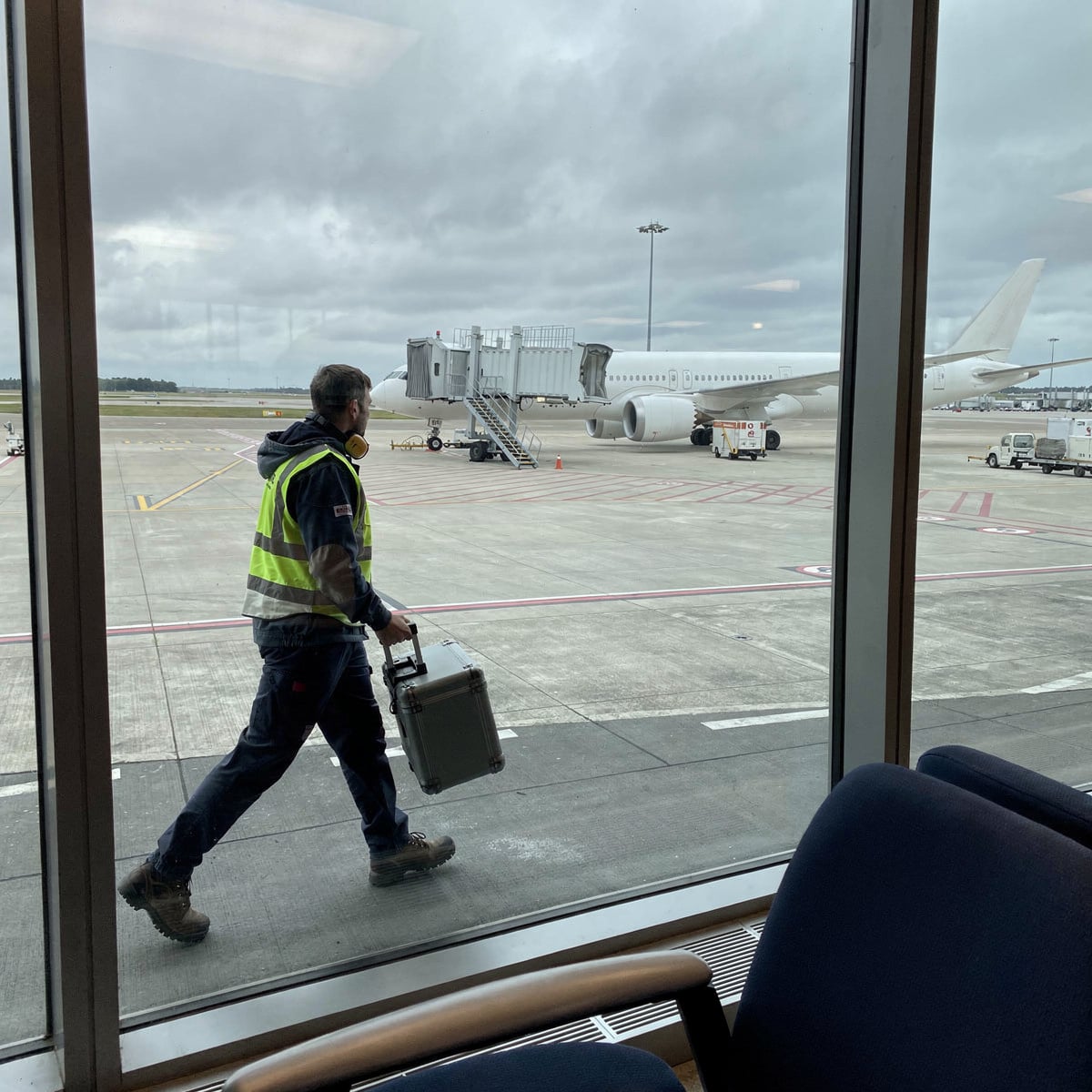 Overcast midday at an international concourse, seen from a seated passenger's position inside the terminal: floor-to-ceiling glazing in the near foreground, and beyond it an open apron where a single wide-body sits at a stand with a jetbridge connected and ground equipment at rest.