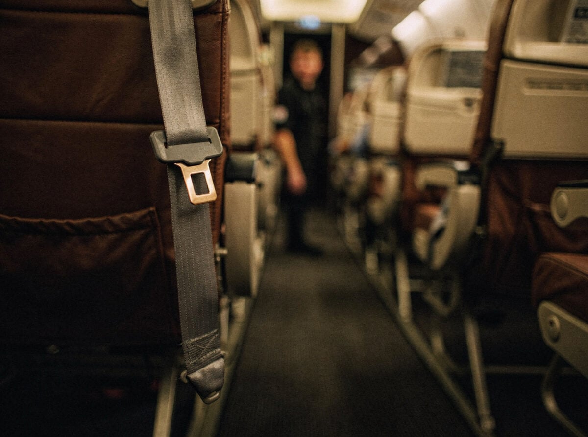 An empty aircraft cabin aisle at night, with a single unbelted seatbelt hanging loosely in the foreground, illuminated by soft amber mood lighting.