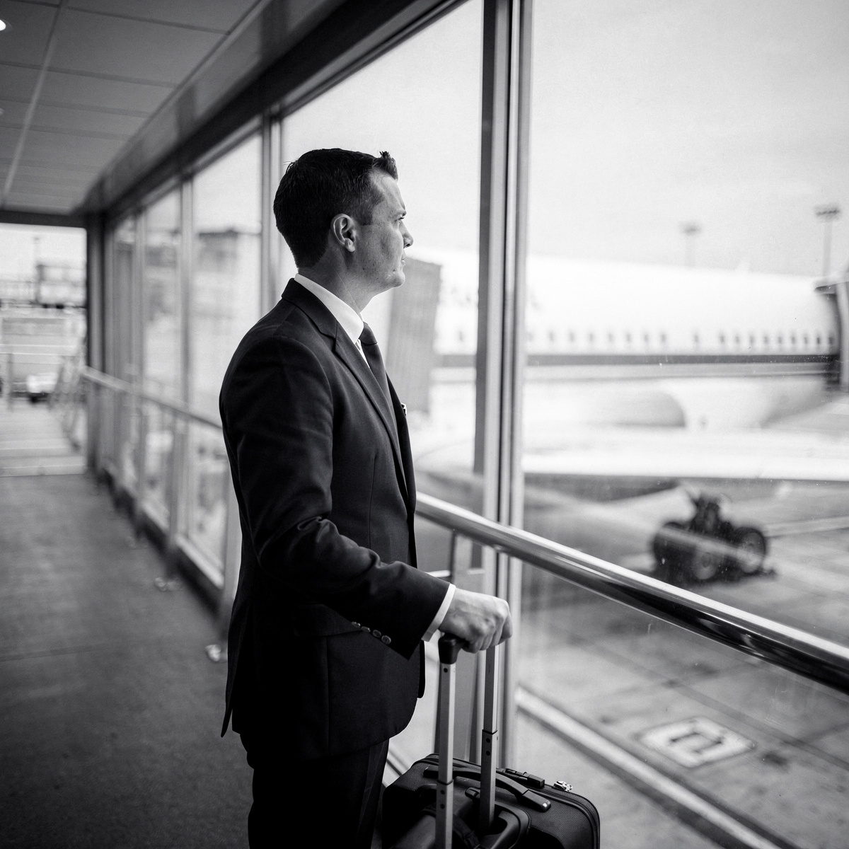 A pilot in a dark uniform stands in an empty, brightly lit jetbridge, looking out at a white aircraft through the glass.