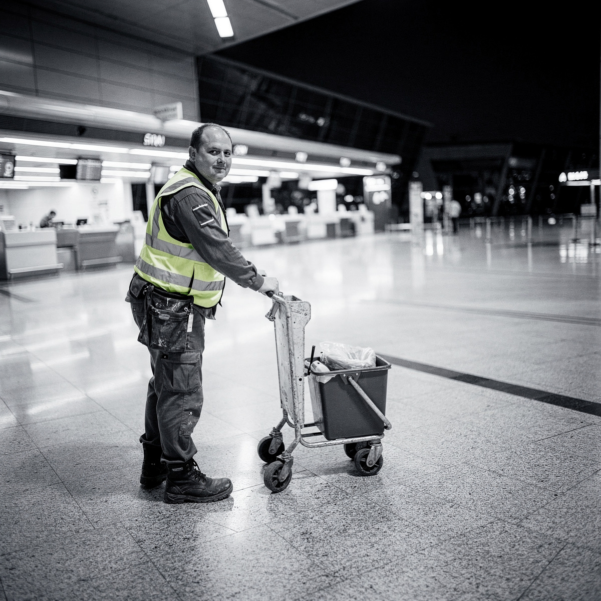 An empty airport terminal at night, bathed in the cool glow of artificial light, with a cleaning crew member working in the foreground.