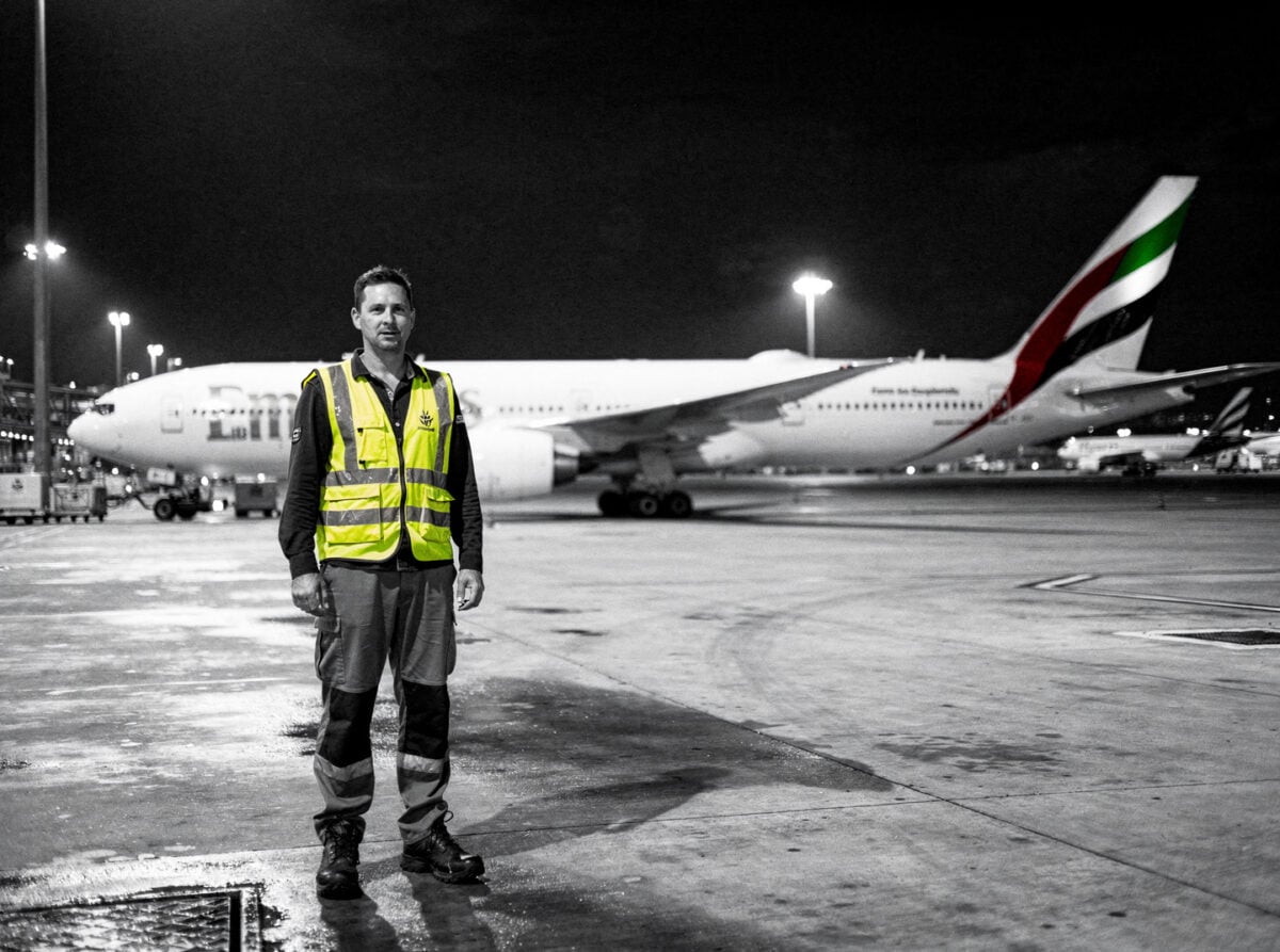 Sydney Airport's apron at night, with an Emirates Boeing 777-300ER parked under floodlights, suggesting a late-night operation during a curfew period.