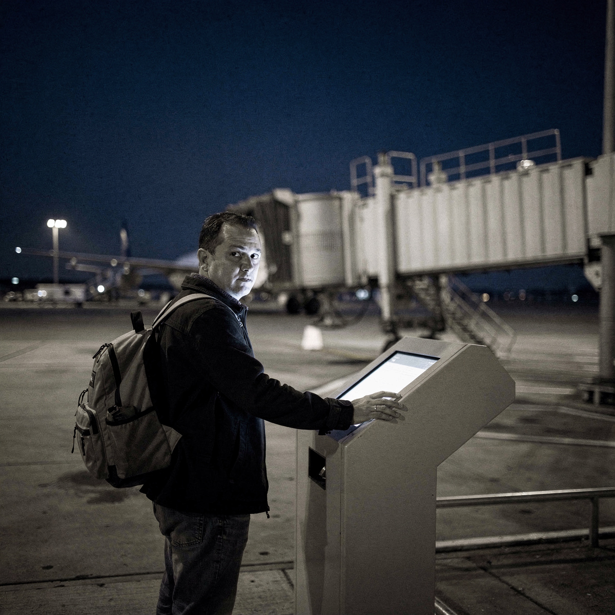 An airport gate area at night, with a single traveler standing near a gate podium, their face illuminated by the soft glow of a screen.