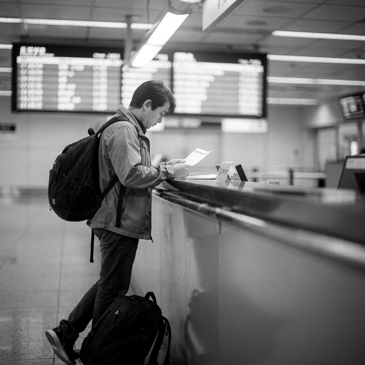 A traveler stands at a counter in a Chinese airport terminal, reviewing documents, with a blurred departure board in the background under harsh overhead fluorescent light.