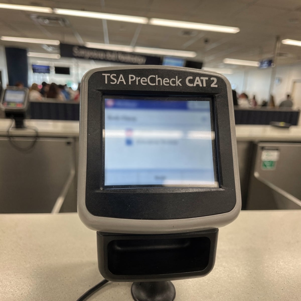 A close-up of a TSA PreCheck CAT-2 unit at an airport security checkpoint, showing the screen with a blurred, unreadable interface. The unit is mounted on a counter, with a generic airport background.