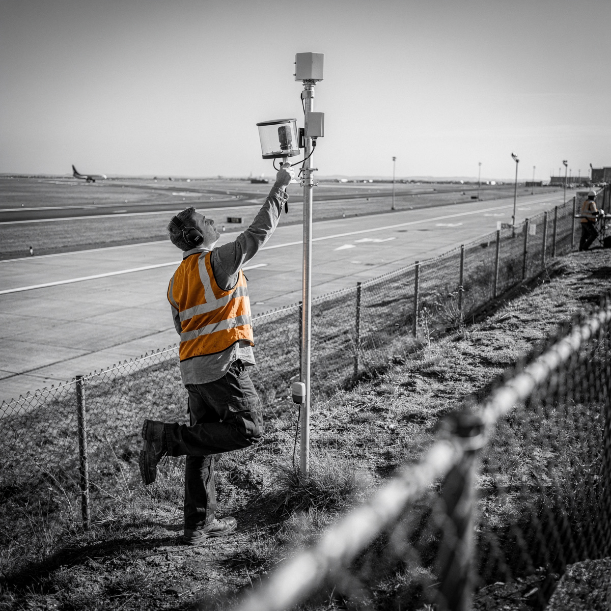 Airport perimeter at midday, a boundary fence line and grass verge seen from an elevated vantage, a slim noise monitoring mast standing beside the fence with a single runway strip and approach lighting receding beyond.