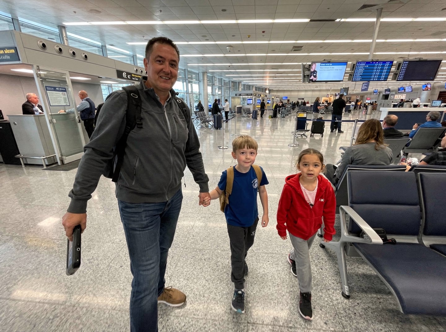 A single parent and two children navigate a busy airport terminal during the day, highlighting the challenges of family seating arrangements.