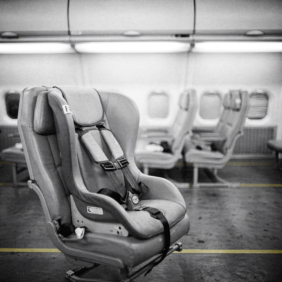 An empty aircraft cabin, cool white overhead lighting, with a child restraint system secured into a passenger seat.