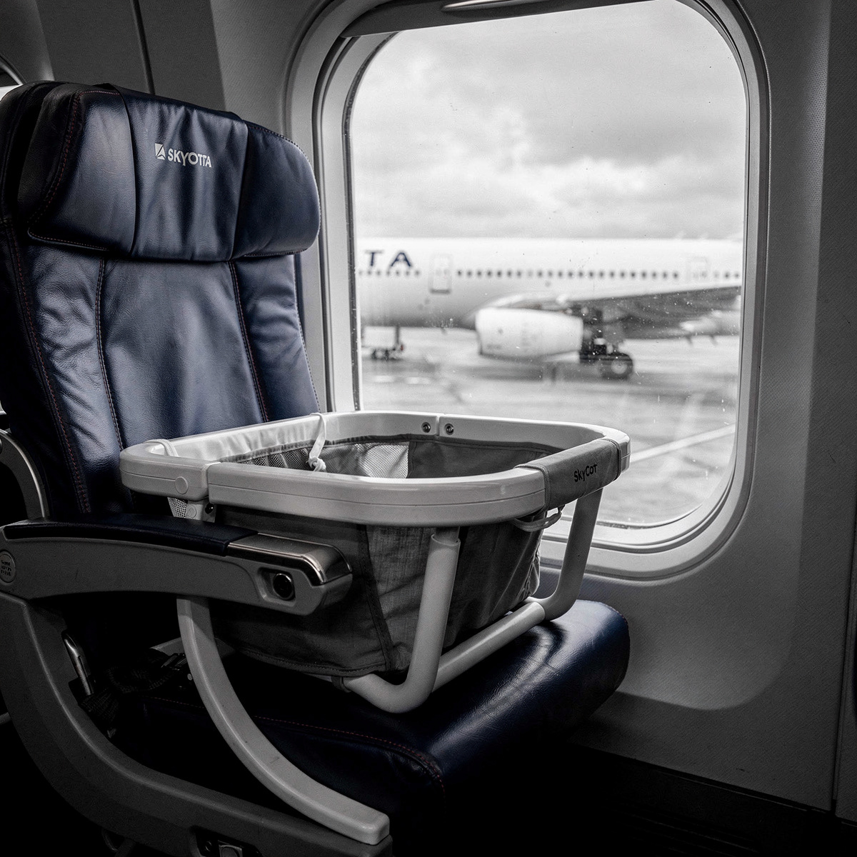 A Delta Air Lines Airbus A330-300 sits at a gate during an overcast day, a SkyCot bassinet resting on a seat inside the cabin, visible through a window.
