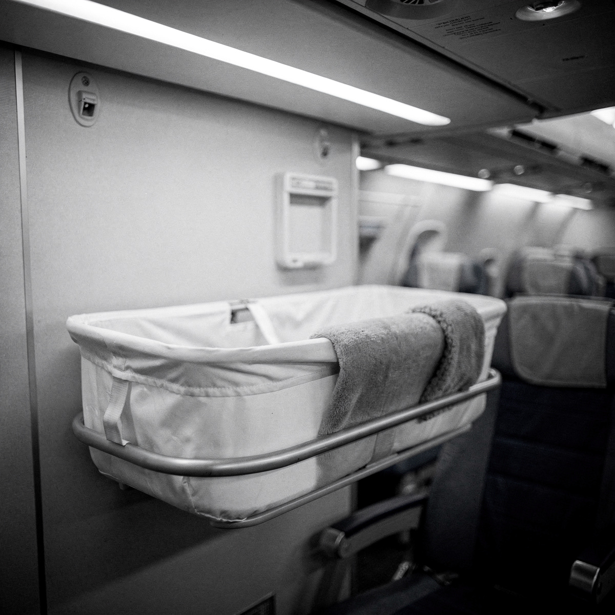 An empty bassinet, neatly prepared, sits on a bulkhead in a commercial aircraft cabin under cool white overhead lighting.