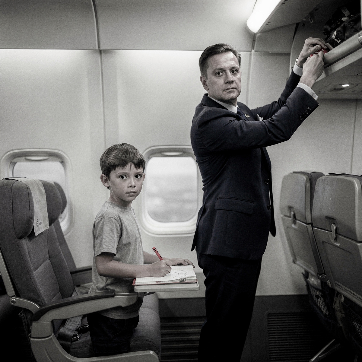 An economy class cabin, mid-flight, with overhead lighting creating a cool, functional atmosphere.