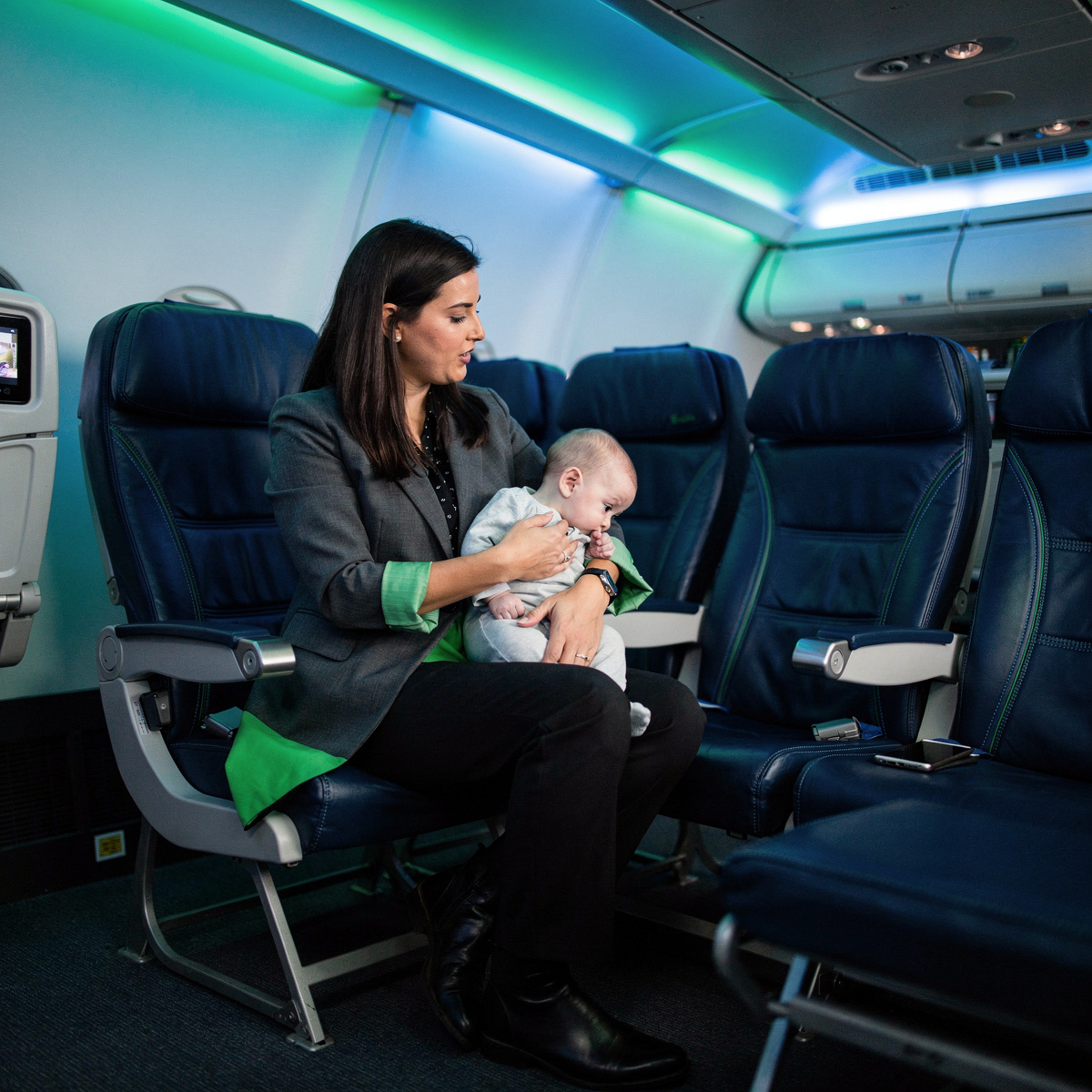 A mother holds her lap infant in a narrow-body aircraft cabin, bathed in soft blue and vibrant green LED mood lighting, during a routine flight.