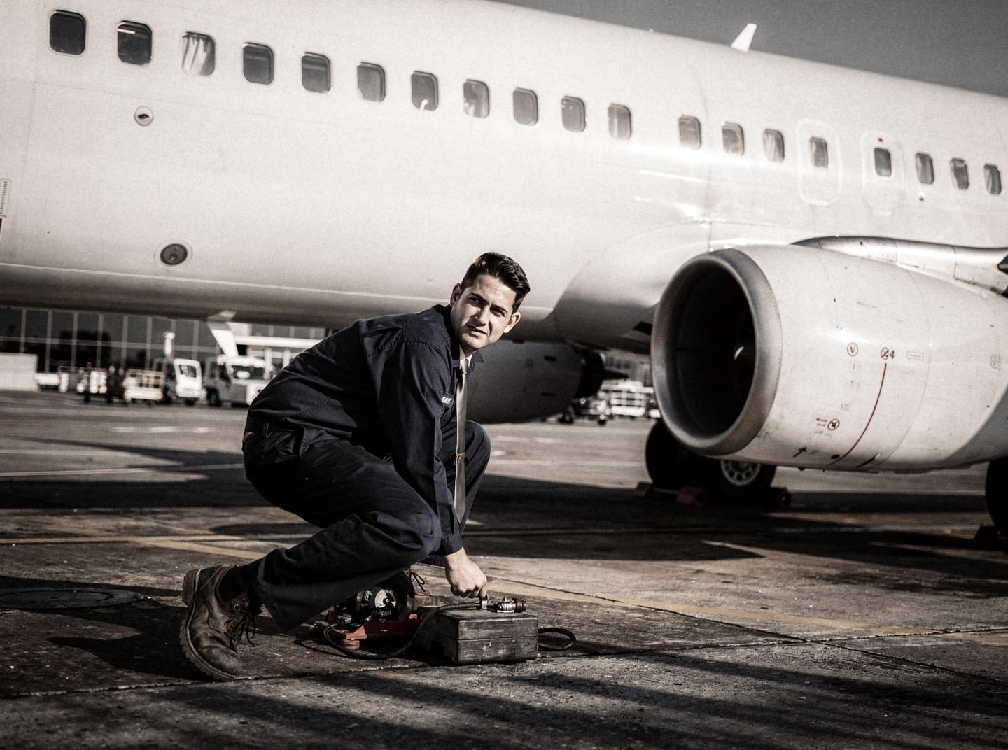 A ground crew member works on a Boeing 737-800 at an airport gate under harsh midday sun.