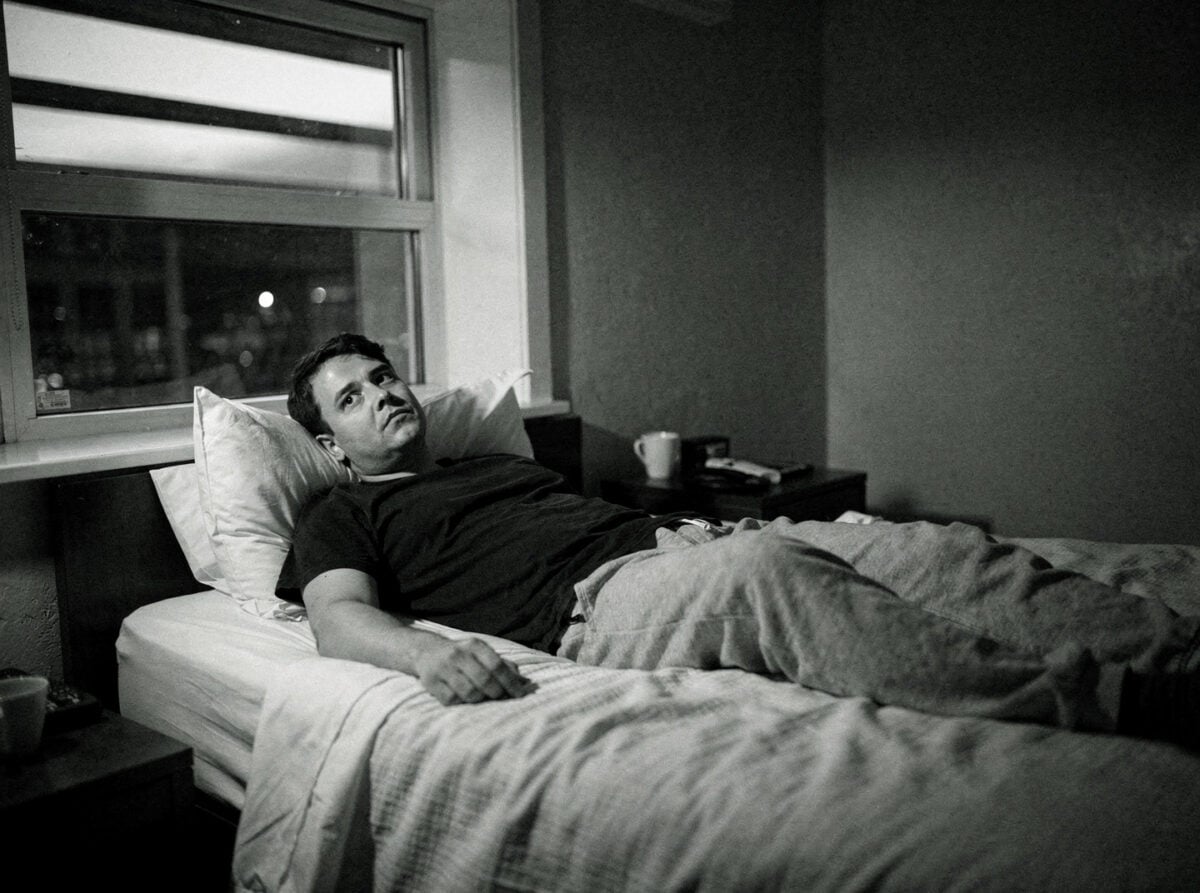 A person lies awake in a darkened hotel room bed during the early hours of the destination night, the room dimly lit by ambient light from a window.