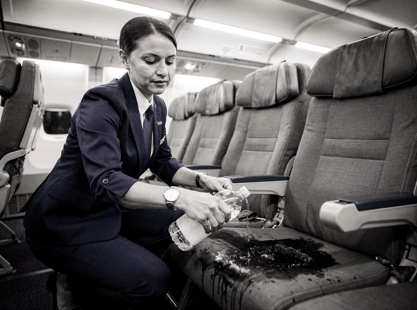 A flight attendant in a generic commercial aircraft cabin, applying water from a bottle to a smoldering seat cushion. The cabin is lit by standard cool white overhead lighting.