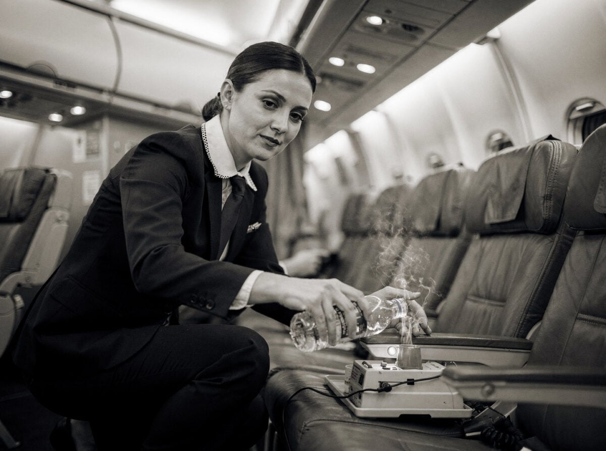 A flight attendant in a standard aircraft cabin, mid-flight, performing a routine safety procedure. The cabin is lit by cool white overhead lights, creating a functional atmosphere.