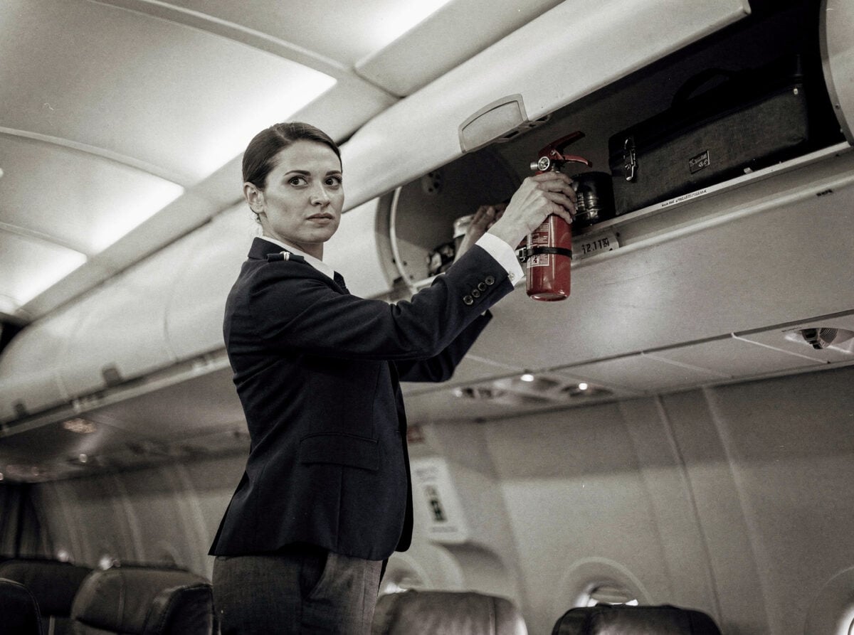 Overhead bin in a commercial aircraft cabin, with a crew member attending to it, during an institutional routine moment in the late afternoon.