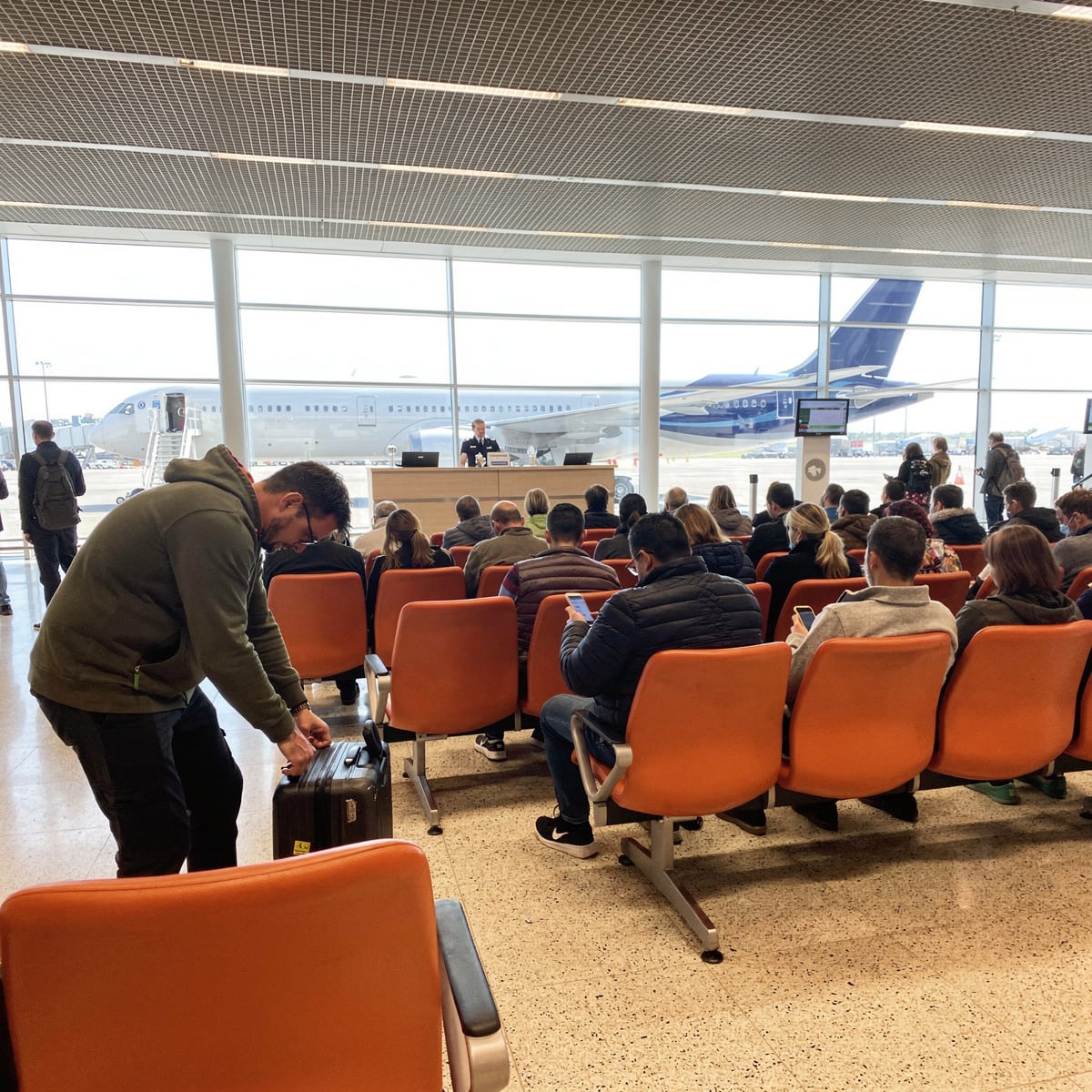 Gate seating area of a modern terminal under harsh midday light, rows of moulded orange plastic seats filled with waiting passengers, a glazed wall beyond showing a partially visible wide-body aircraft at its stand.