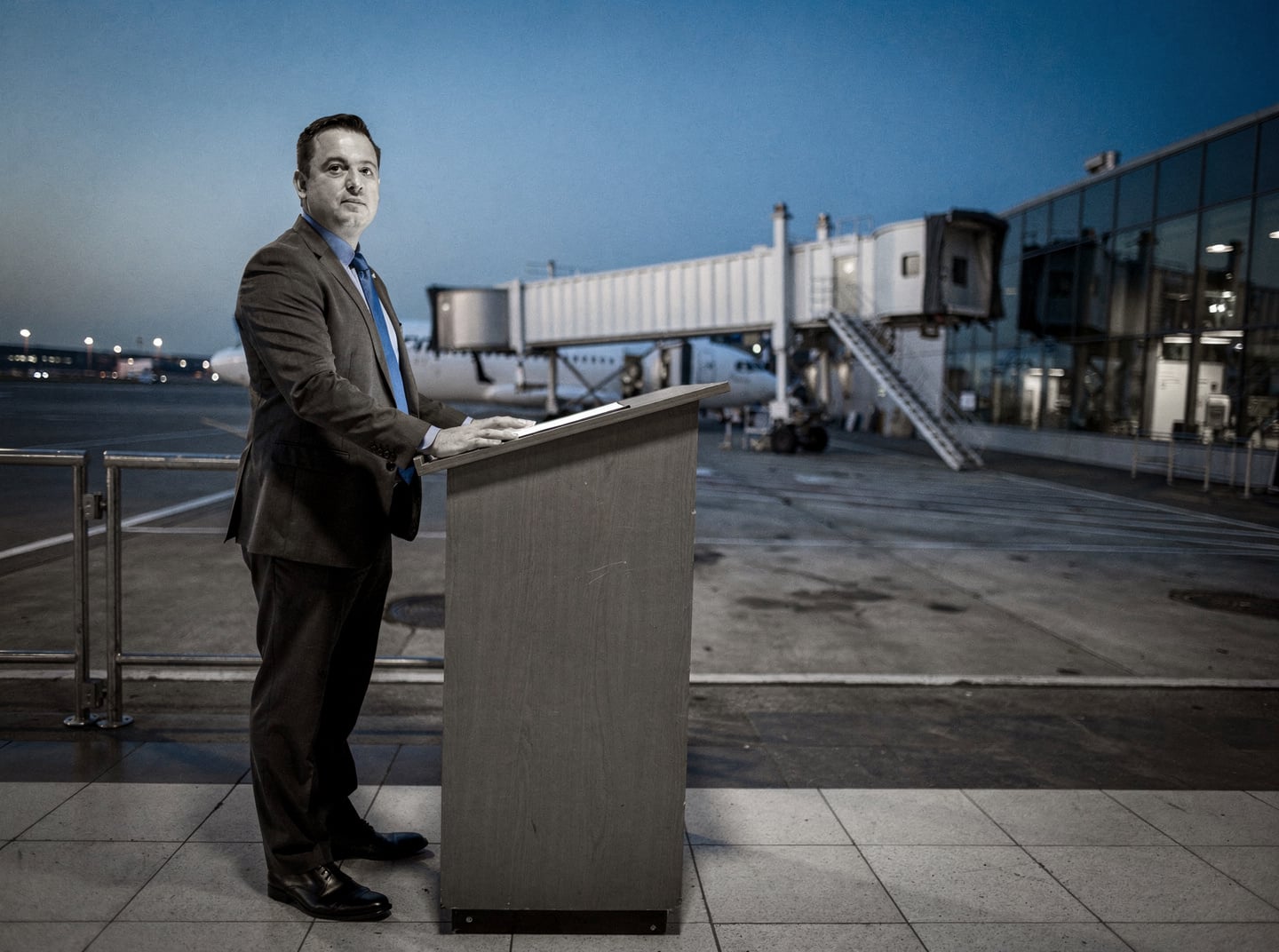 A single worker stands at a gate podium in a quiet airport terminal during blue hour, with an empty jetbridge visible beyond.
