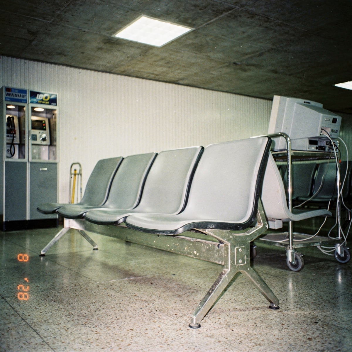 Empty 1980s airport transit lounge interior at midday, boxy concrete ceiling, rows of molded plastic seating, a CRT monitor turned away on a steel trolley, no people.