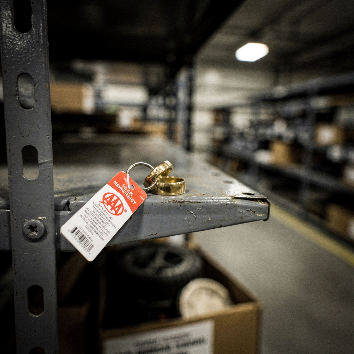 A wedding ring on a keyring with a AAA membership tag sits on a metal inventory shelf in a dimly lit government surplus warehouse, surrounded by other confiscated items.