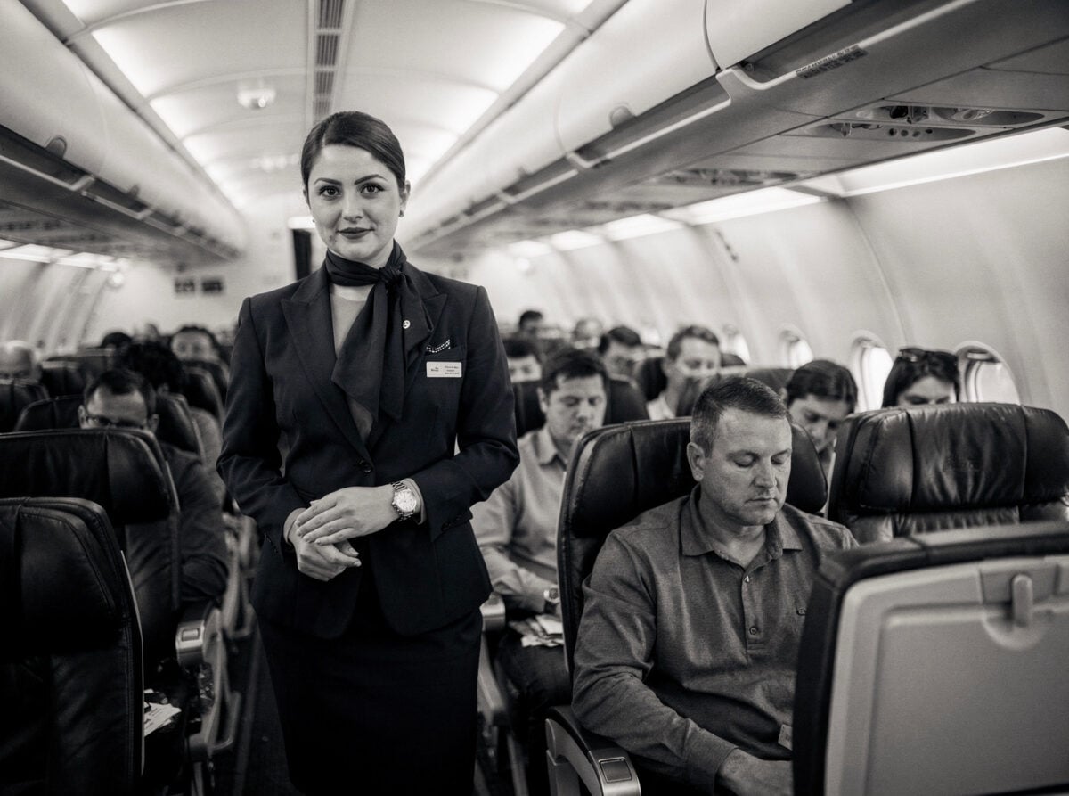 An aircraft cabin with passengers seated, viewed from a slightly elevated position, during a gate hold or taxi with all doors closed.