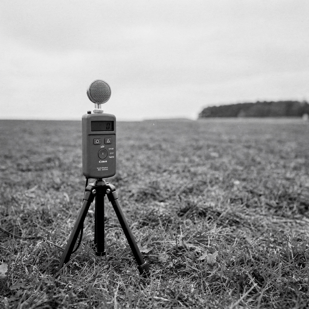 A sound measurement device, mounted on a small tripod, stands on a grassy field under an overcast sky, ready to record ambient noise levels.